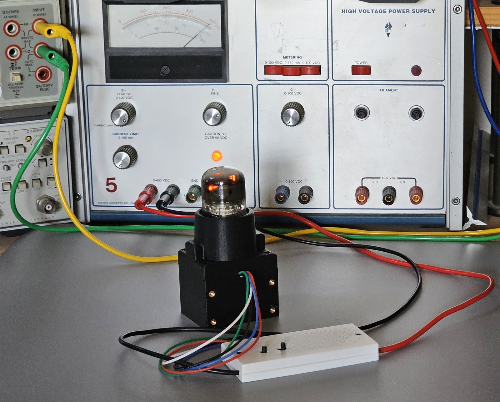 The A101 bench fixture running: the tube lit in its printed socket housing, orange glow inside the glass, in front of the high-voltage bench supply, with the white clamshell holding the bench circuit and its two pushbuttons in the foreground