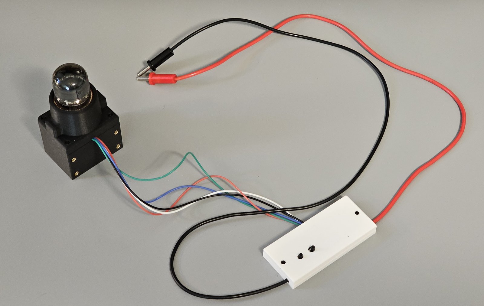 The assembled A101 bench fixture: the tube in its black socket housing and shroud, wired to the white clamshell with two pushbuttons on top, and the red and black HV leads